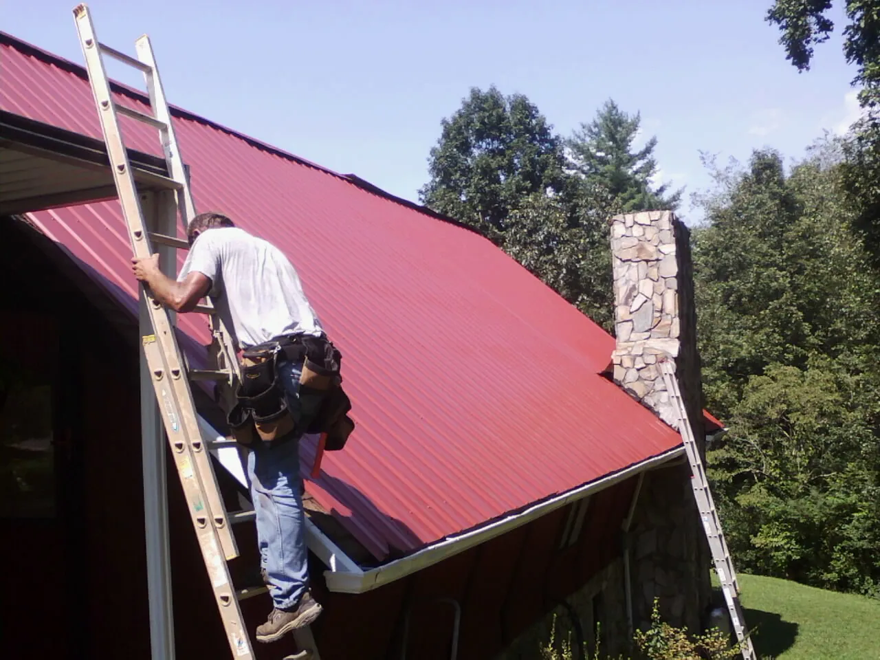 Roofer on a ladder beside a red metal roof and stone chimney — Soffit & Fascia Repair by Roofing Brothers in Cooperstown